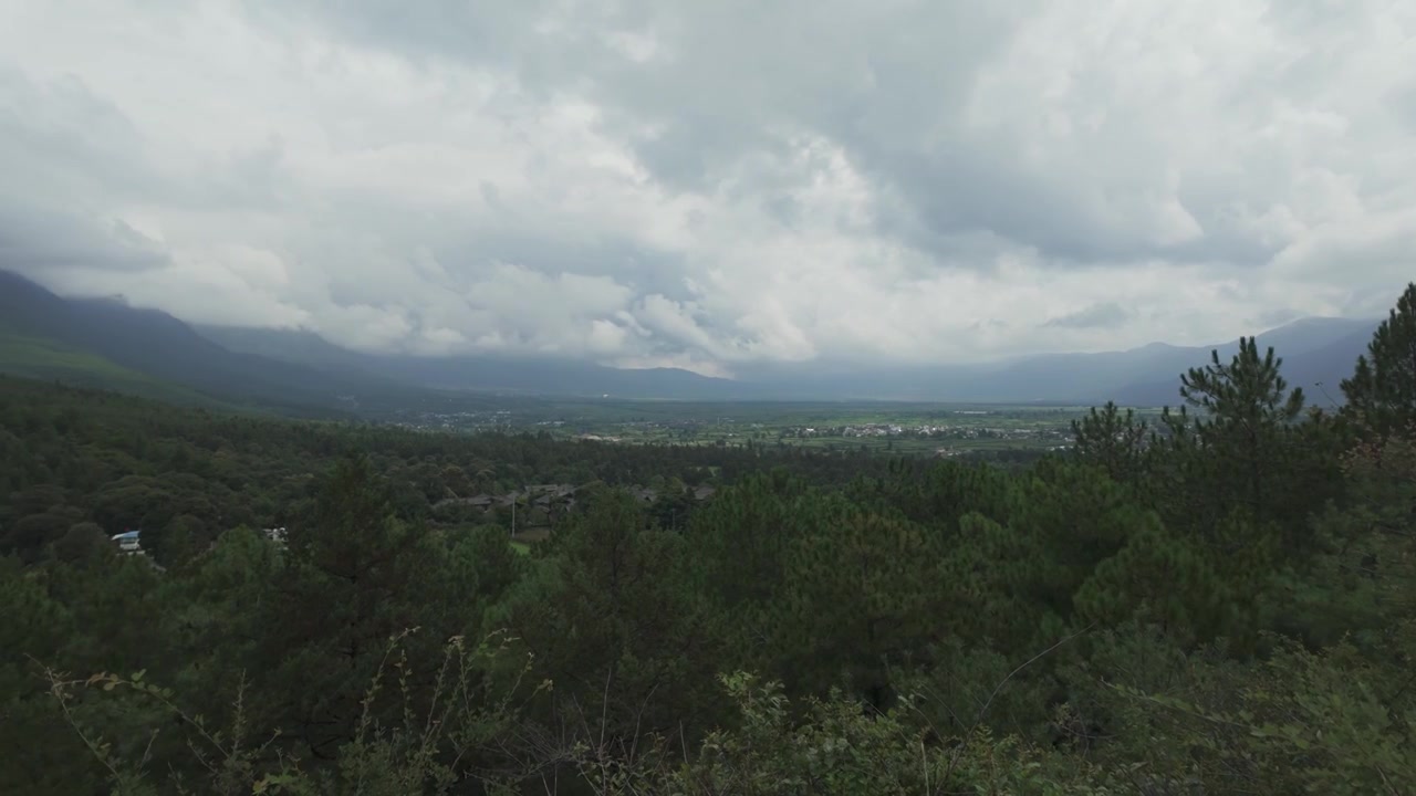 A cinematic wide shot overlooking the lush green valley of Baisha Town and Fuguo Monastery, nestled beneath a dramatic overcast sky in Yunnan.