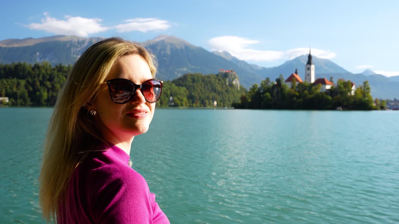 Smiling female blonde tourist on lake shore looking out toward Bled Island