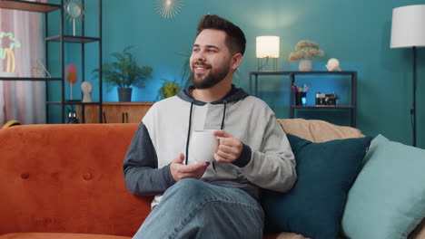 attractive smiling caucasian man drinking a cup of coffee or herbal tea at home couch in the morning