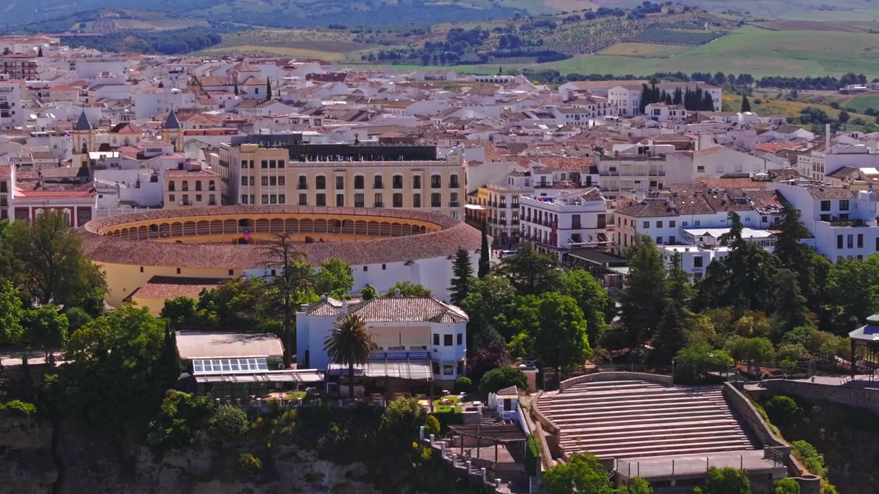 Riprese drone che si allontanano e si inclinano verso l'alto oltre l'anfiteatro di Ronda, i giardini terrazzati e il campanile della chiesa, svelando lentamente l'intera città imbiancata a calce e la catena montuosa sfocata oltre