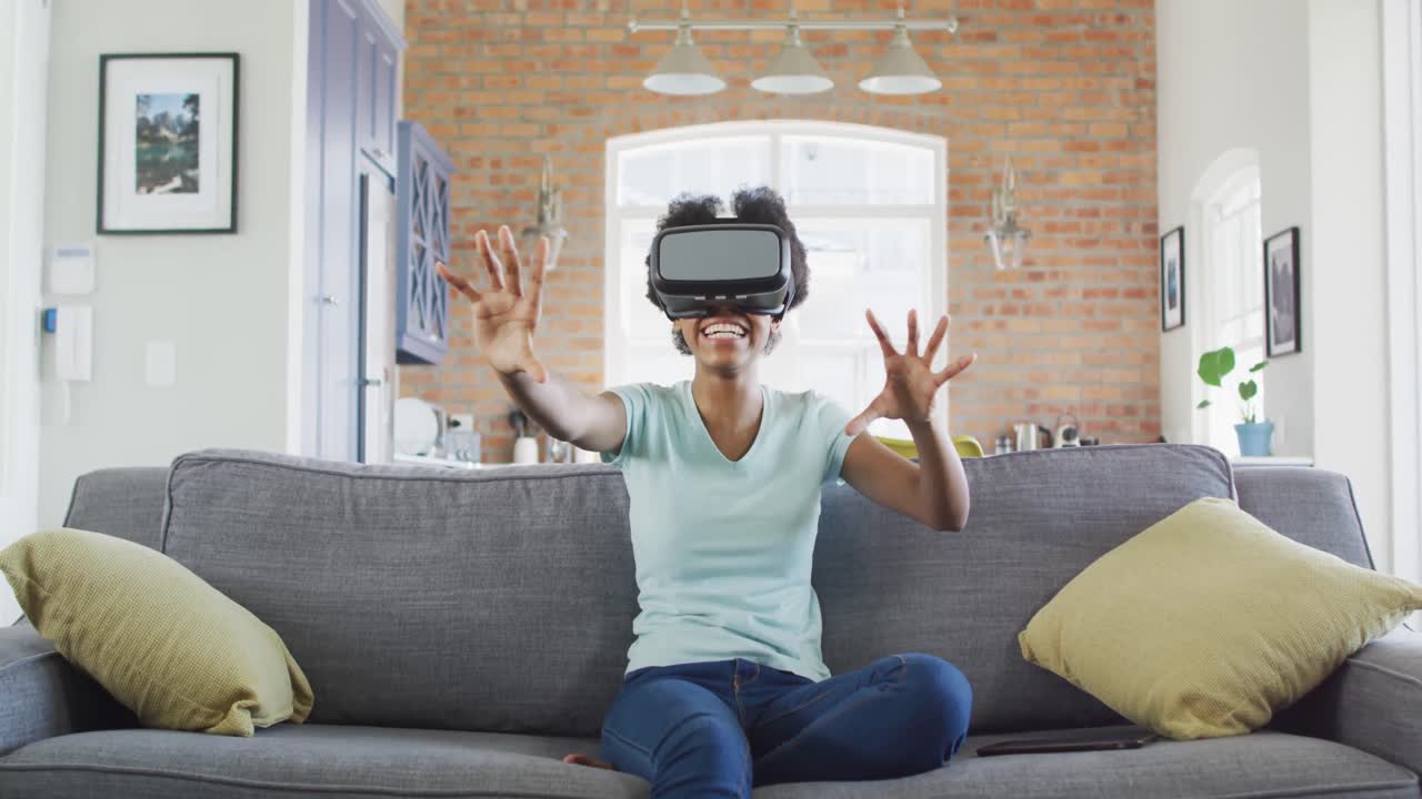 Happy african american teenage girl sitting on sofa, wearing vr headset and touching virtual screen