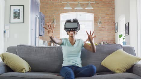 feliz adolescente afroamericana sentada en el sofá, con auriculares vr y tocando la pantalla virtual