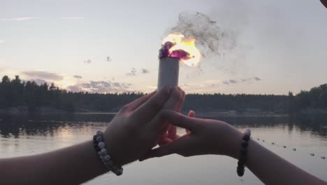 Dos-Personas-Sostienen-Una-Bomba-De-Humo-Coloreada-En-Las-Manos-Mientras-Caminan-Hacia-Un-Lago,-Cámara-Lenta