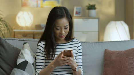 Young-Woman-In-Striped-Blouse-Tapping-And-Texting-A-Message-On-The-Smartphone-Sitting-On-Sofa-In-The-Living-Room-1