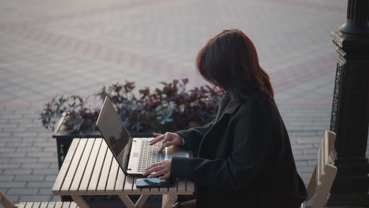 Young lady typing on laptop outdoors with hair cascading down, hand reflection visible on phone screen on wooden table, background features decorative flower arrangement and pillar in urban