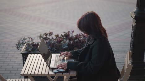 joven escribiendo en una computadora portátil al aire libre con el cabello en cascada hacia abajo, el reflejo de la mano visible en la pantalla del teléfono en la mesa de madera, el fondo presenta arreglos florales decorativos y pilares en la ciudad