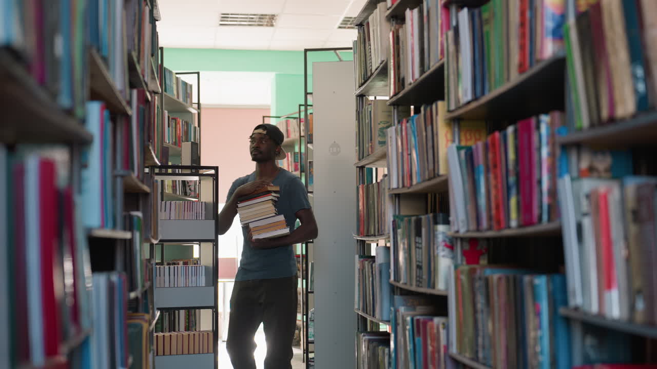 Young man in casual t-shirt and cap walks through narrow library aisle holding books, turning around toward shelves filled with colorful books under bright indoor lighting