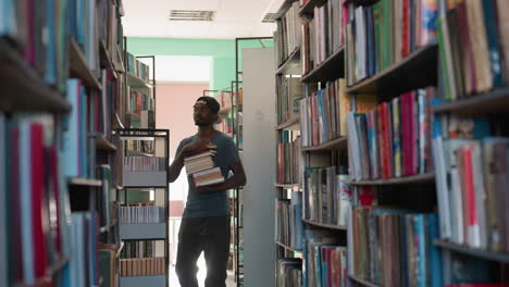 young man in casual t-shirt and cap walks through narrow library aisle holding books, turning around toward shelves filled with colorful books under bright indoor lighting