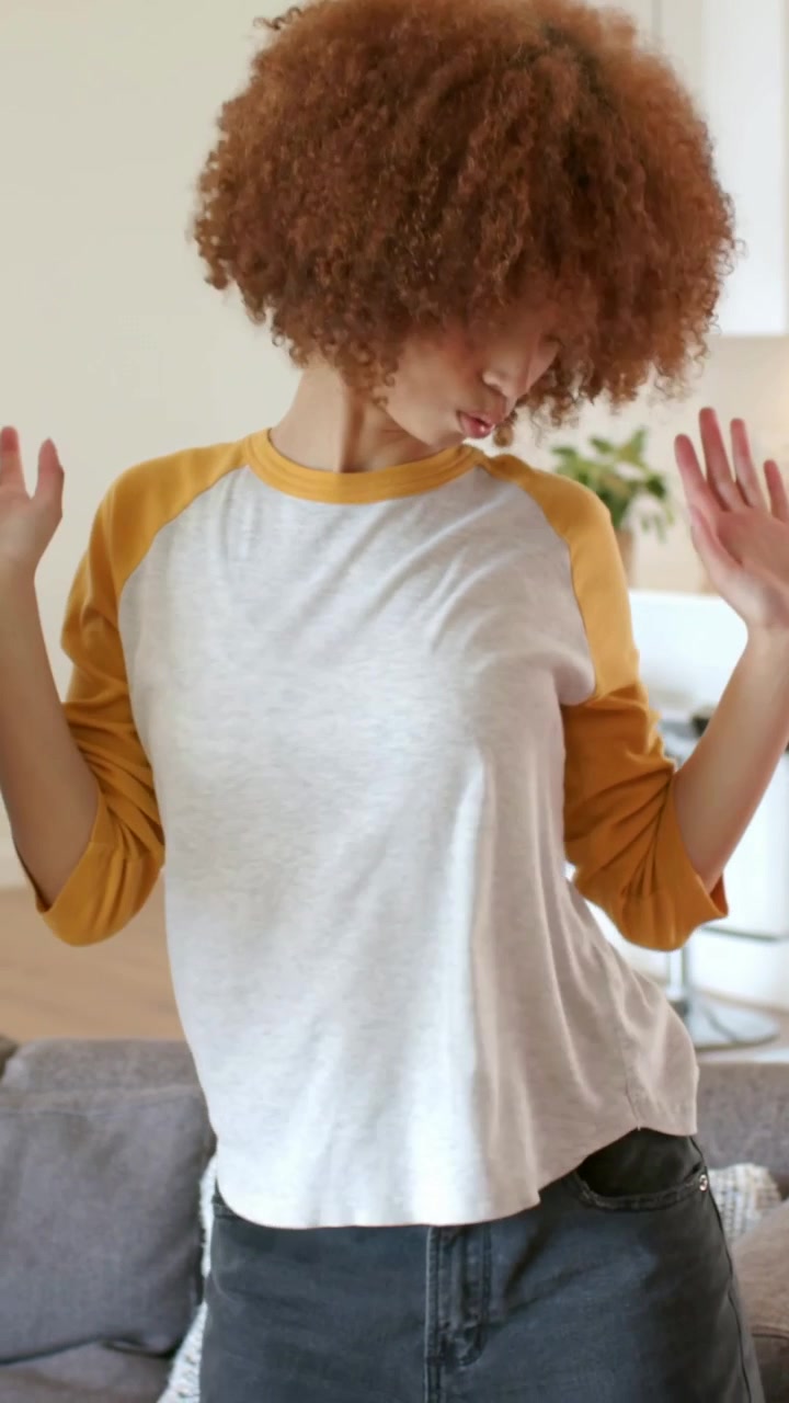 Vertical video: Dancing woman with mustard sleeves responding to off-camera arm near sofa at home