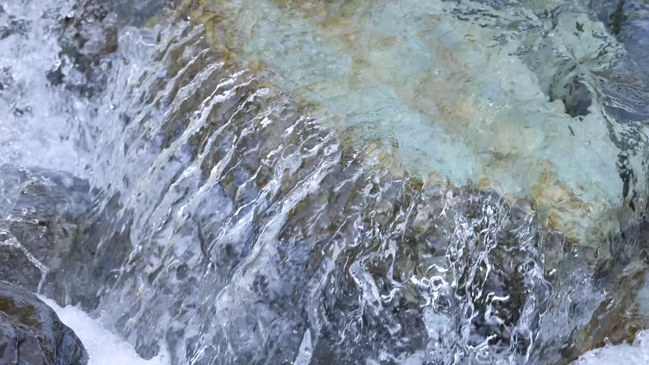 Crystal-clear water cascades over moss-covered rocks in a natural mountain stream, captured in daylight with a steady, close-up camera angle