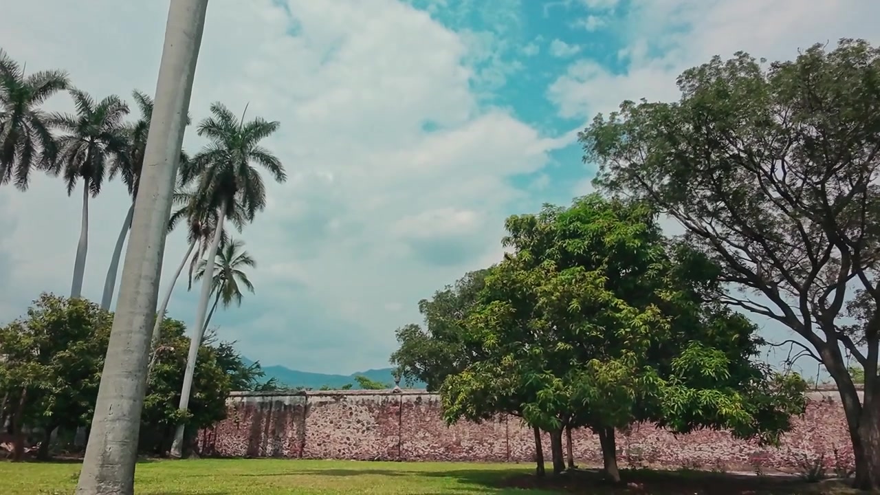 Alte palme e giardini paesaggistici accanto al muro di una storica hacienda a Morelos, Messico, con alberi secolari, montagne distanti e cielo blu parzialmente nuvoloso.