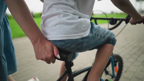 a close-up a woman s hand holding the seat of a bicycle as her son rides along a paved walkway