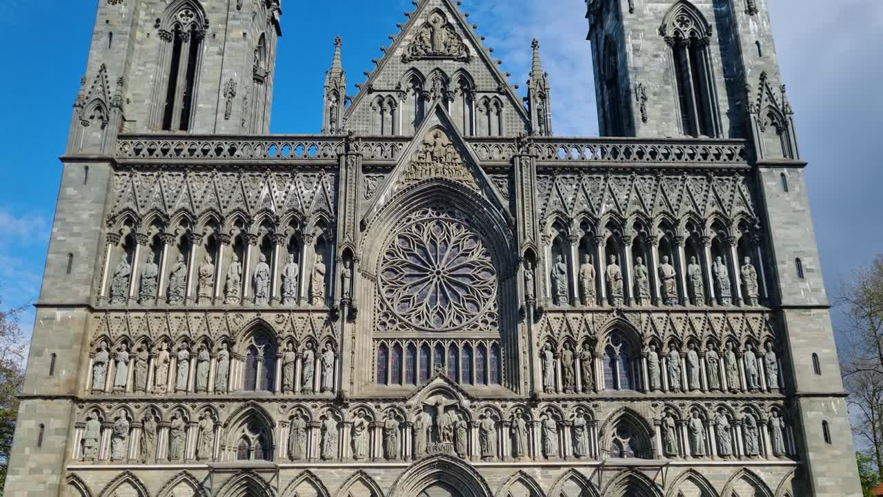 Revealing shot of the front entrance of Nidaros Cathedral in Trondheim, Norway, under a clear blue spring sky, showcasing its intricate Gothic architecture, detailed stone carvings and pointed arches