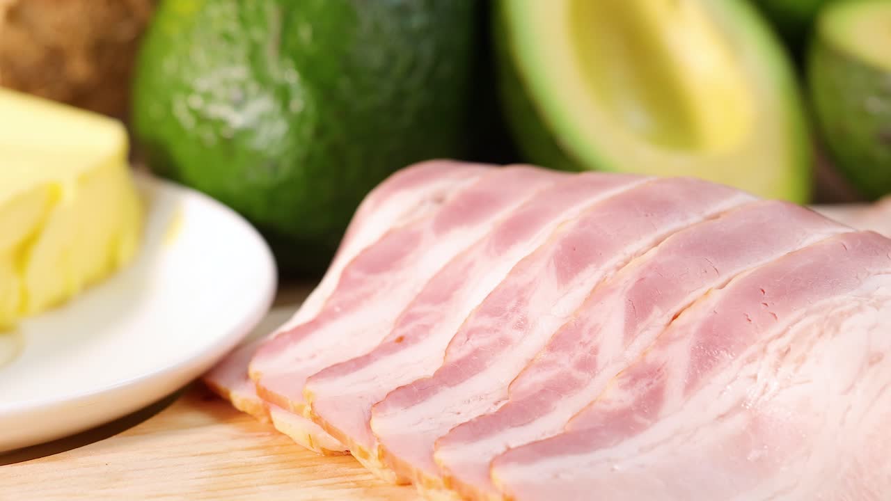 A vibrant arrangement of avocados, bacon, butter, seeds, and nuts under bright lighting on a wooden surface