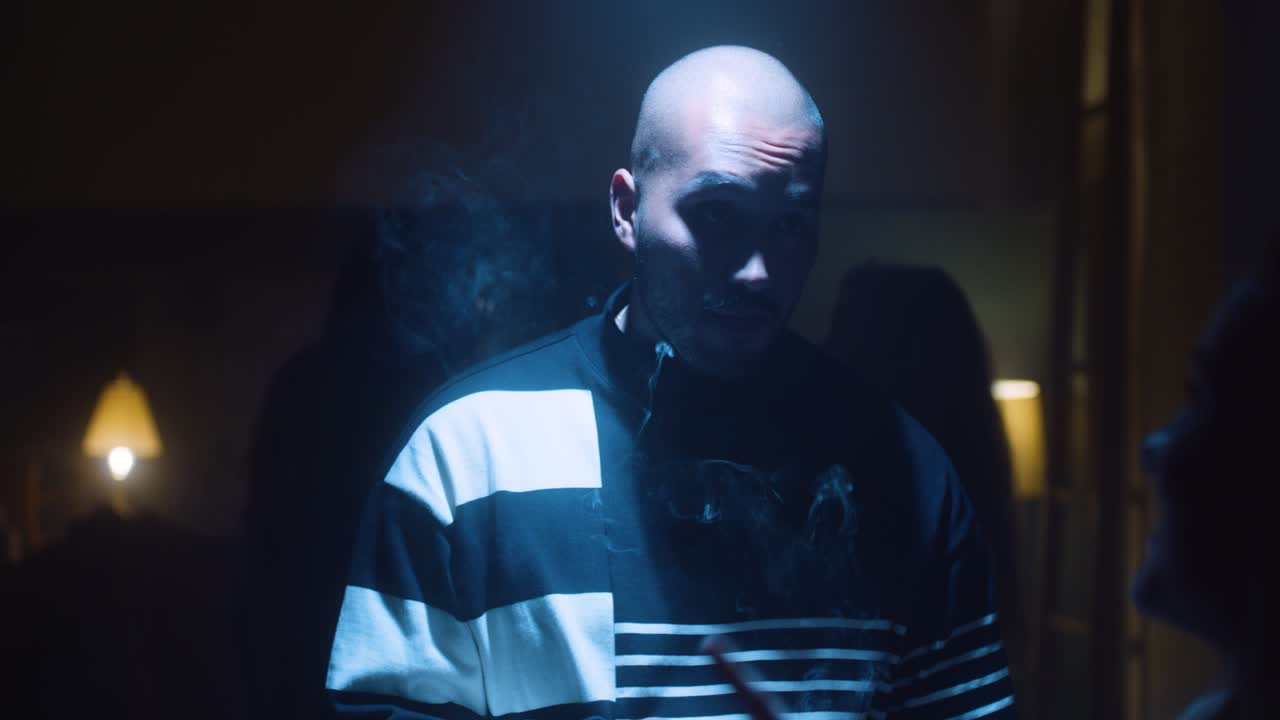 Man smokes, spot lit at a party while an angry girl walks up.