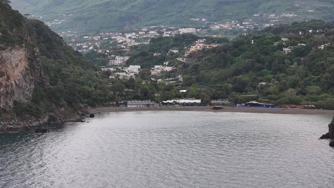 bahía rocosa con playa de arena y ciudad de montaña en la isla de ischia, italia, avión no tripulado
