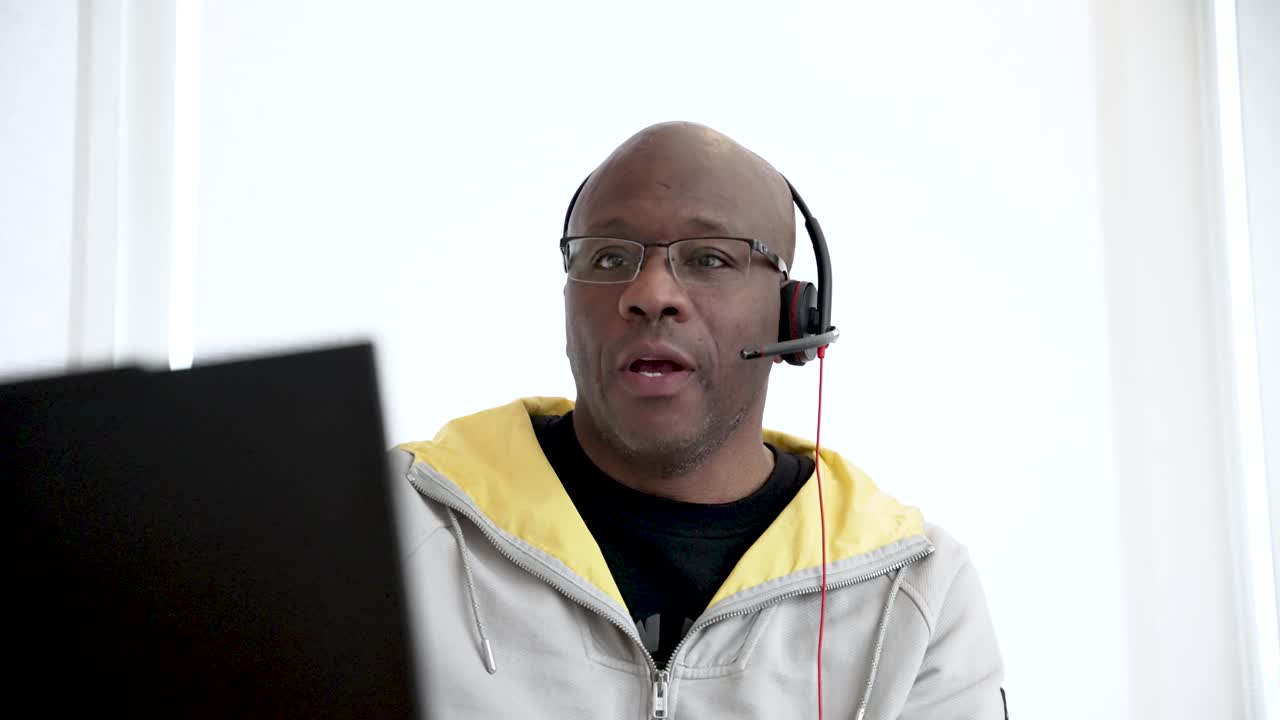 Black male man wearing a headset working remotely on a laptop in casual attire.