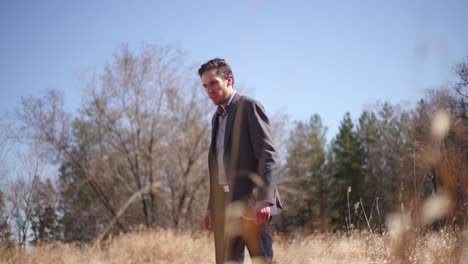 Man-in-suit-walking-across-a-meadow-filled-with-tall-grass-and-surrounded-by-trees-while-contemplating-his-surroundings