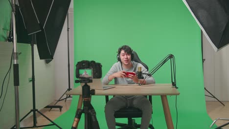 una cámara grabando a un jugador asiático con auriculares y computadora agitando la mano mientras juega un juego en un teléfono móvil en pantalla verde con equipo de luz profesional