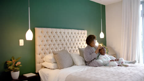 Happy-african-american-mother-combing-daughter-hair-sitting-on-bed-in-bedroom,-slow-motion