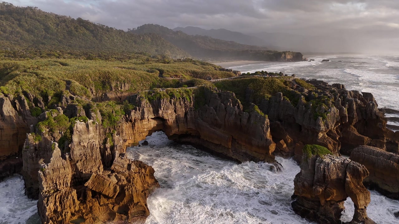 Ocean waves surging through a natural arch in the Pancake Rocks at sunset in Punakaiki, New Zealand. Aerial drone