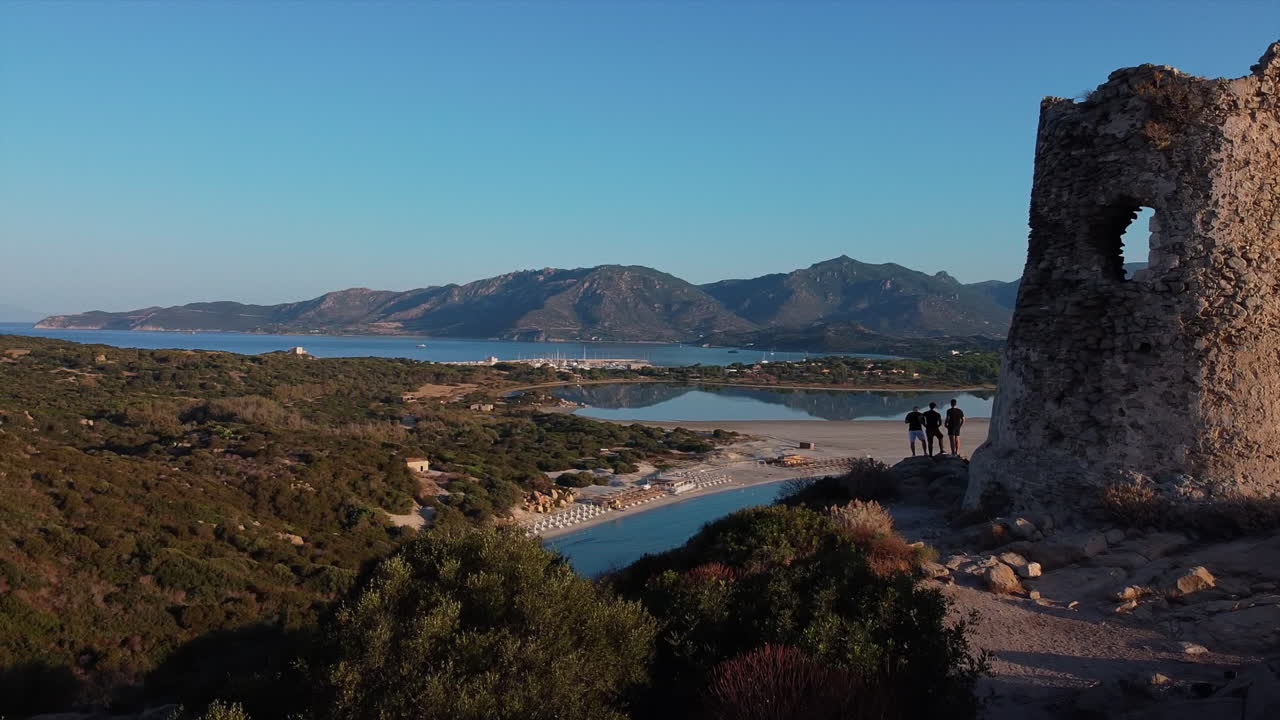 vista de drones de un grupo de tres amigos varones parados en la torre de porto giunco mirando a través del hermoso mar, las playas y la montaña durante el día en italia