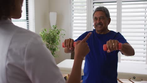 Fisioterapeuta-Femenina-De-Raza-Mixta-Ayudando-A-Un-Hombre-Mayor-A-Hacer-Ejercicio-Con-Pesas