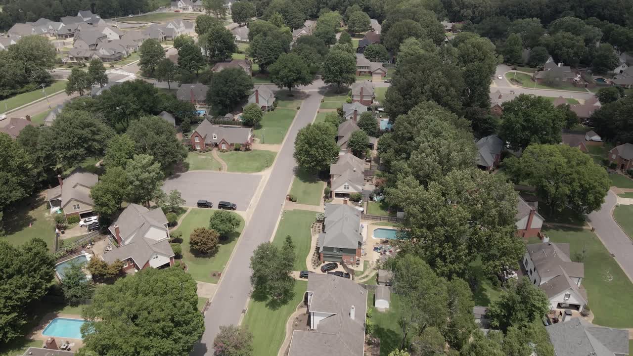 A drone zoom-in shot capturing a suburban neighborhood in the United States, showcasing tree-lined streets and residential houses. The greenery and -planned layout highlight the peaceful