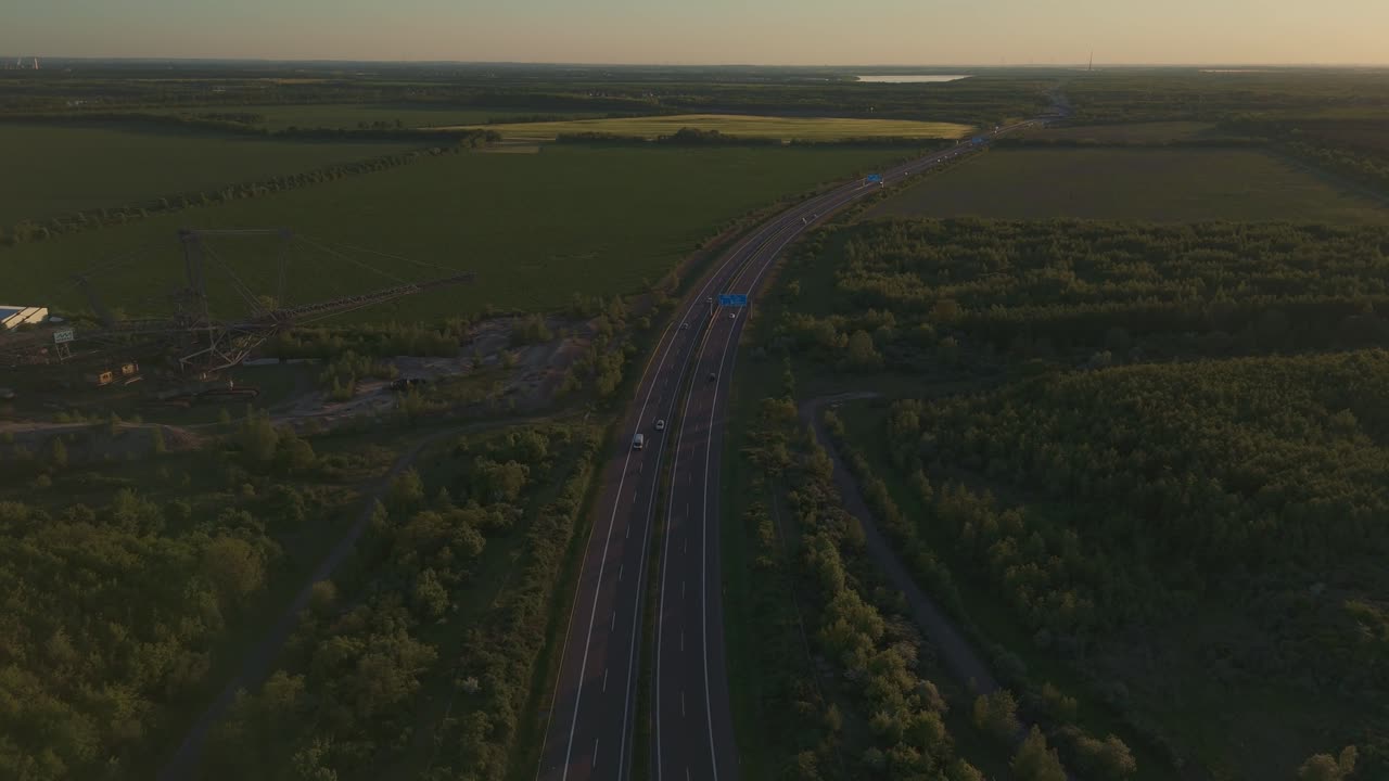 Wide aerial shot of a German autobahn and massive coal mining excavator near Leipzig during sunset. The scene captures the industrial contrast against natural fields and fading daylight.
