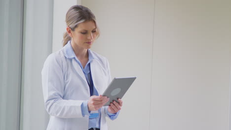 female doctor wearing white coat standing in hospital corridor looking at digital tablet