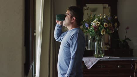 Un-Hombre-Caucásico-Pensativo-Parado-En-La-Ventana-Y-Tomando-Café-En-Casa