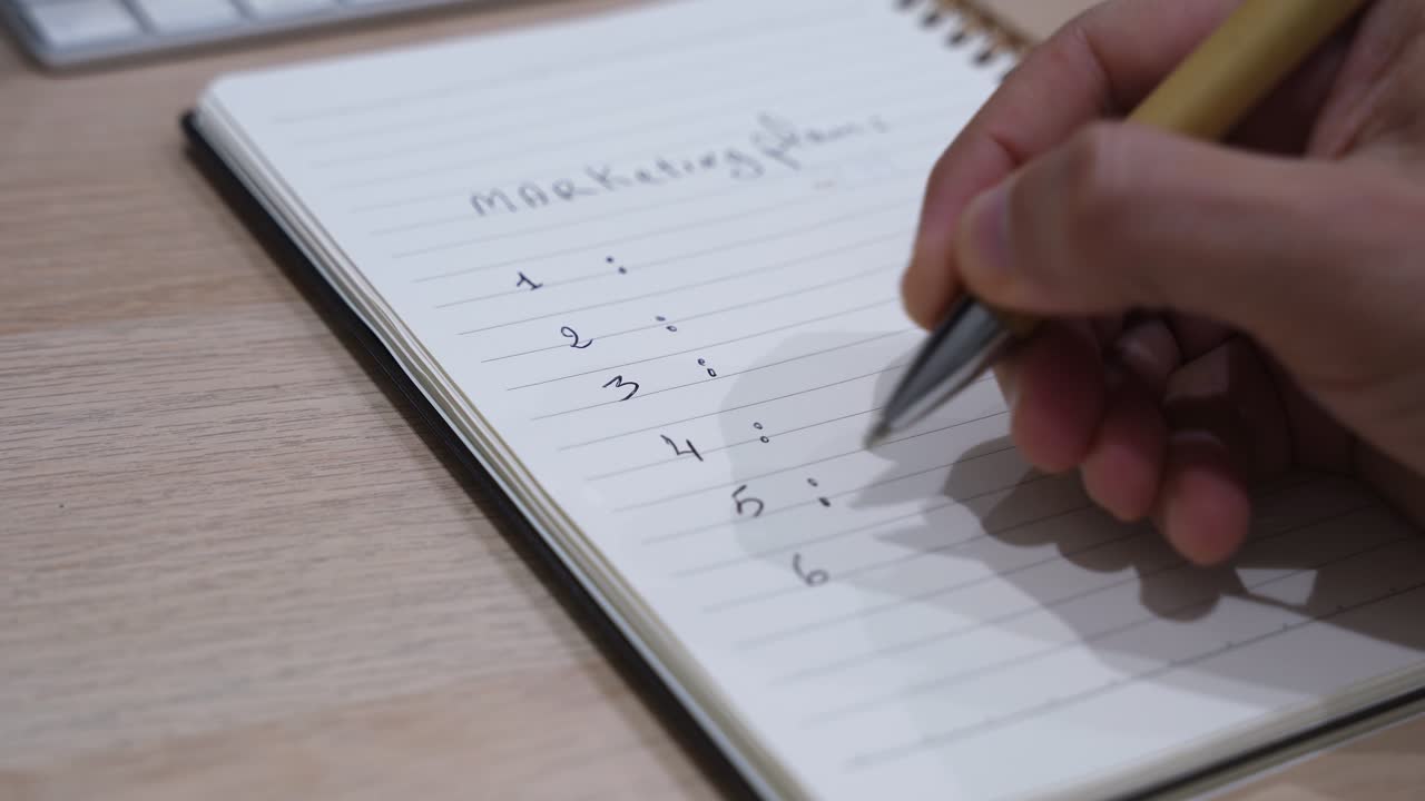 Nærbillede af en hånd, der skriver nummererede trin til en markedsføringsplan i en spiralnotesbog med en kuglepen på et skrivebord, hvilket repræsenterer forretningsplanlægning, strategiudvikling og idéorganisering på en arbejdsplads.