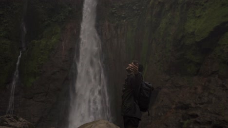 A man with a backpack stands with his arms outstretched, facing a powerful waterfall in the background