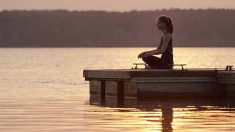 young woman meditating in lotus position on jetty at lake at sunset