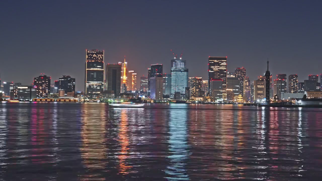 Wide static time-lapse of Tokyo's glittering night skyline across the bay, iconic red tower centered, multicolored city lights reflecting in long vertical streaks on calm dark water.