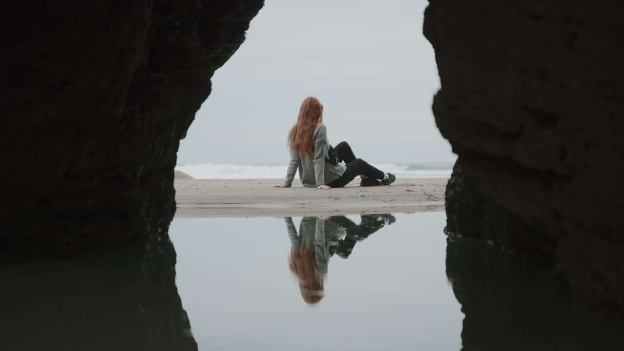 Woman sitting on a beach framed by a sea cave with reflection