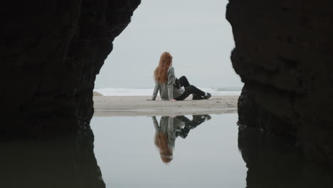 woman sitting on a beach framed by a sea cave with reflection