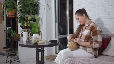 kind hearted woman seated on sofa selecting card from basket on lap in cozy plant filled living room with white brick wall and round glass table wearing checkered shirt and white skirt