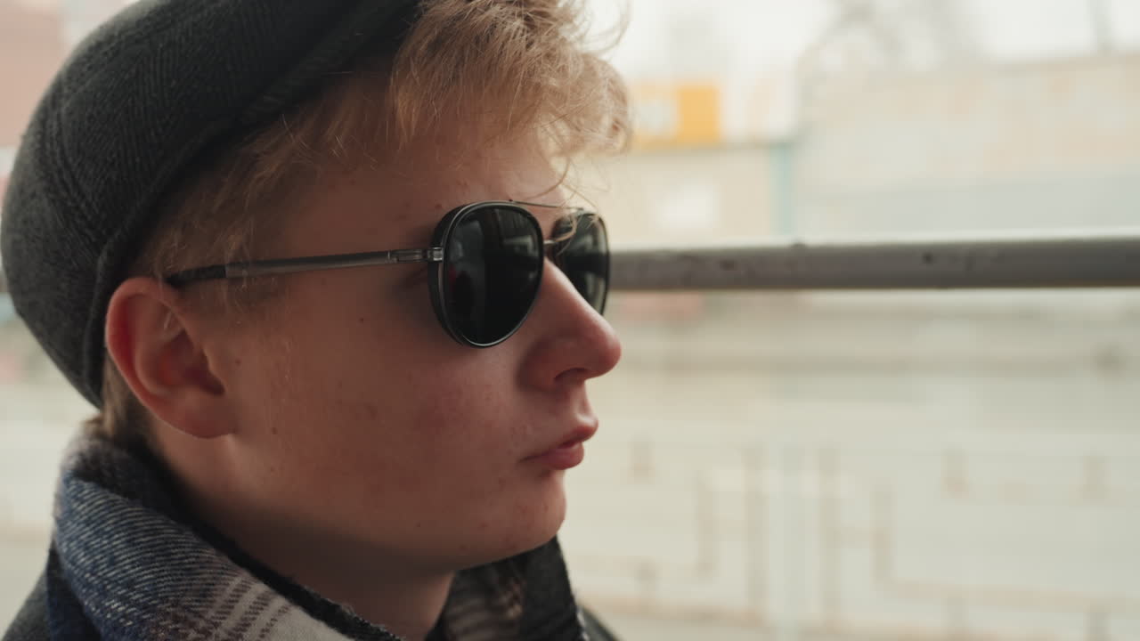Young white creator adjusts sunglasses on cold urban platform, dons flat cap and dark round lenses while gripping metal rail and fixing steady gaze frames suggest director, model, and traveler