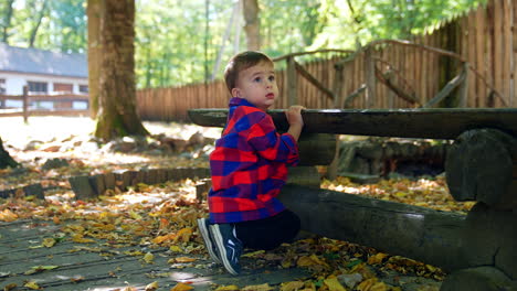 small caucasian toddler boy squatted near the wooden bench. baby sits outdoors on sunny day.