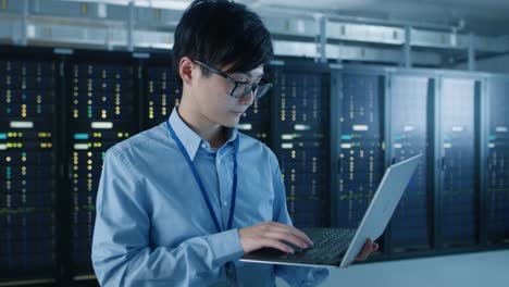 in the modern data center: portrait of it engineer standing with server racks behind him, doing maintenance and diagnostics procedure using laptop. arc camera shot.