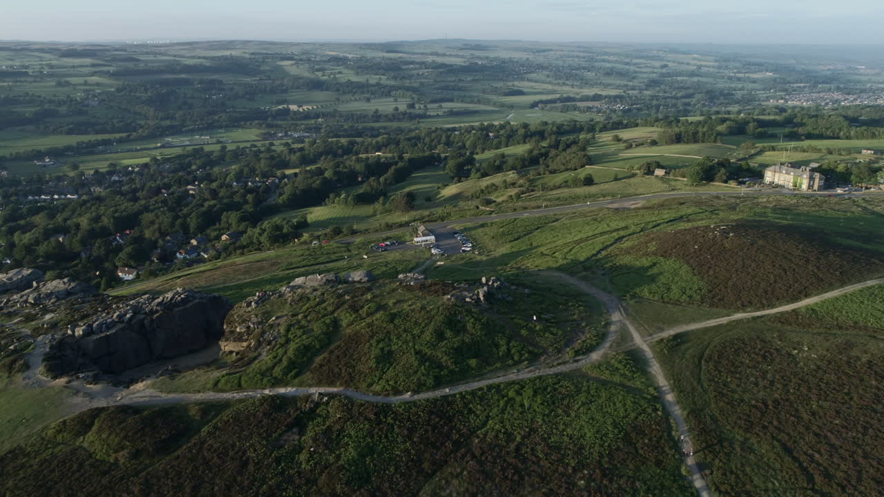 서부 요크셔 도로를 따라 운전하는 자동차와 함께 골든 아워에 와피데일의 벌리를 향해 들판과 나무가 있는 시골 계곡 아래 일클리의 소와 송아지 바위 위로 날아가는 드론 샷을 설정합니다.