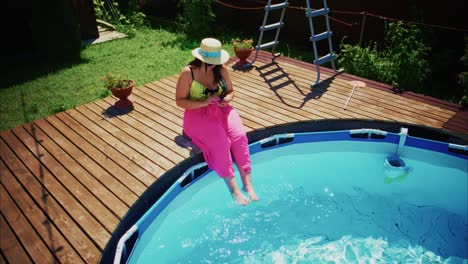 woman in pink pants relaxing by a swimming pool with clear water and wooden deck in sunny weather