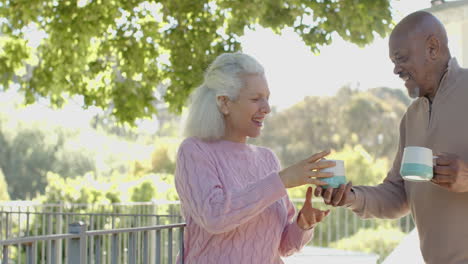 Happy-senior-biracial-couple-embracing-and-drinking-coffee-at-balcony-at-home,-slow-motion