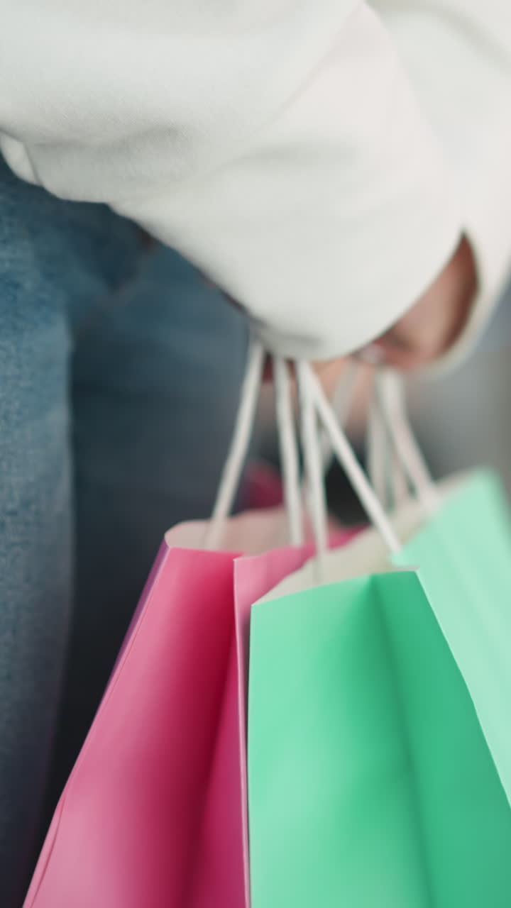 색 스웨트 셔츠와 청바지를 입은 여성이 집 안에 서서 민트와 분홍색 쇼핑 가방을 들고, 가방 손잡이 주위에 미묘한 손 움직임을 하며, 흐릿한 쇼핑몰 배경으로