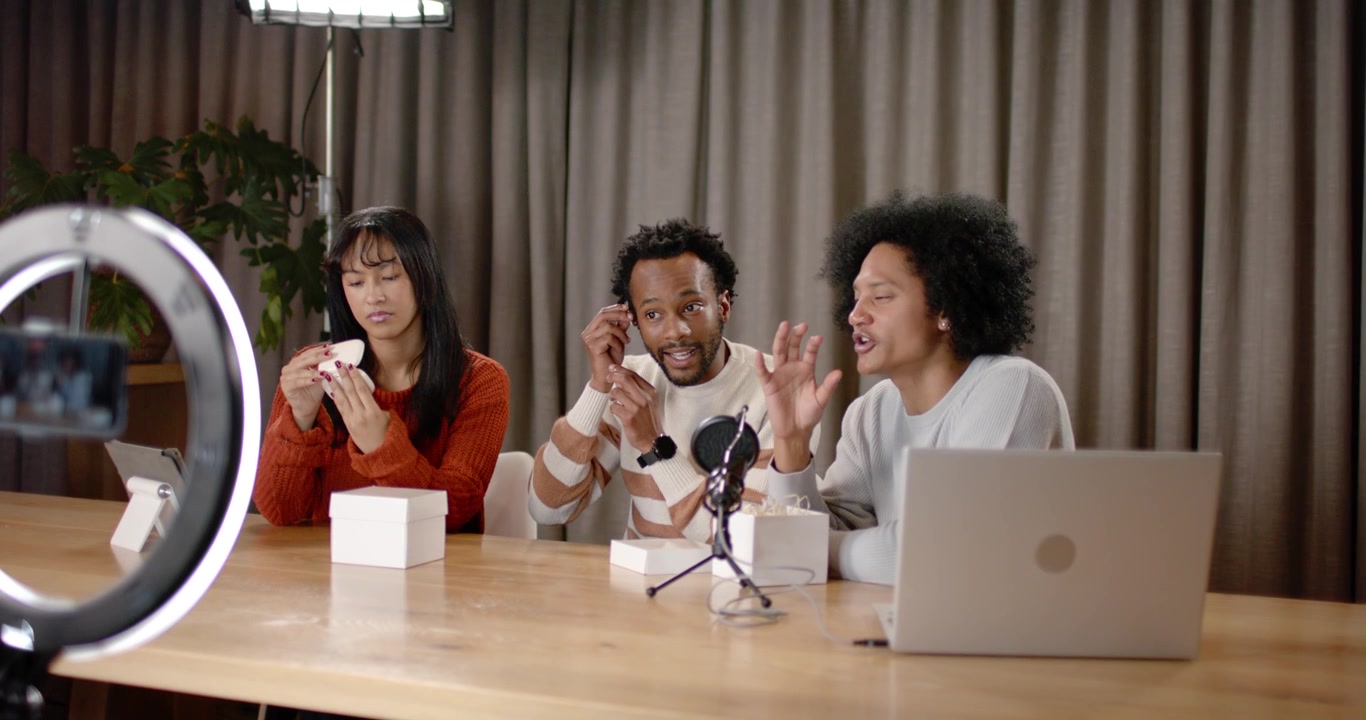 Co-hosts unboxing earbuds after camera prompt, inspecting charging case, testing fit for review