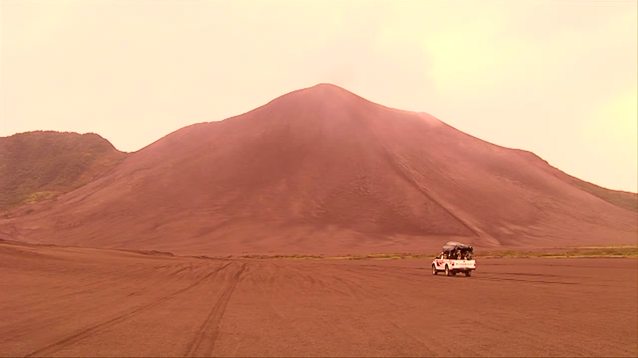 автомобиль движется по пустынному ландшафту, на заднем плане формируется облако вулканического пепла