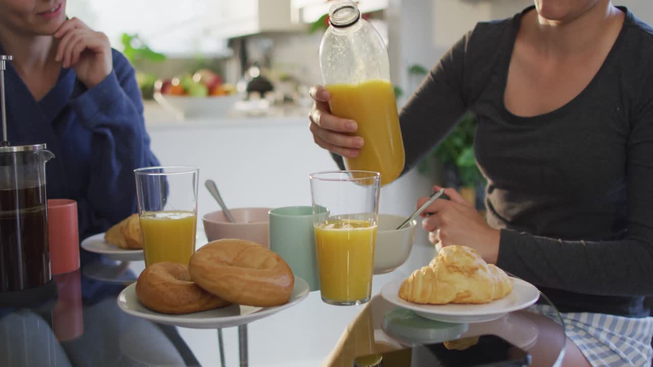 mujer caucásica vertiendo jugo en un vaso mientras desayuna con su esposa en casa