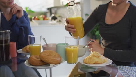 mujer caucásica vertiendo jugo en un vaso mientras desayuna con su esposa en casa