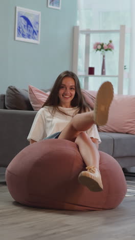 alegre mujer morena en falda gris cae en la silla de bolsa de frijol rosa riendo. la dama disfruta relajándose en la elegante sala de estar después de la universidad